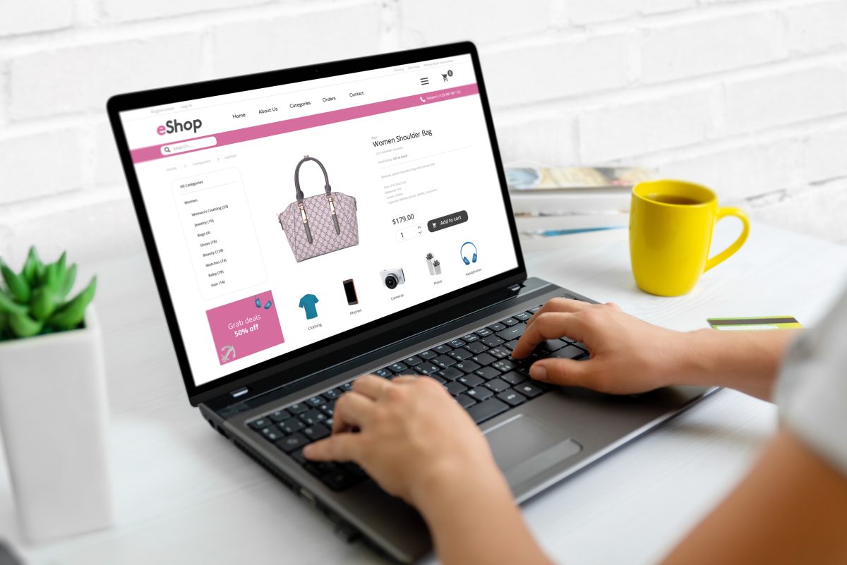 En person sitter vid en bärbar dator med en webbutik öppen på skärmen, med kaffekopp och krukväxt på skrivbordet