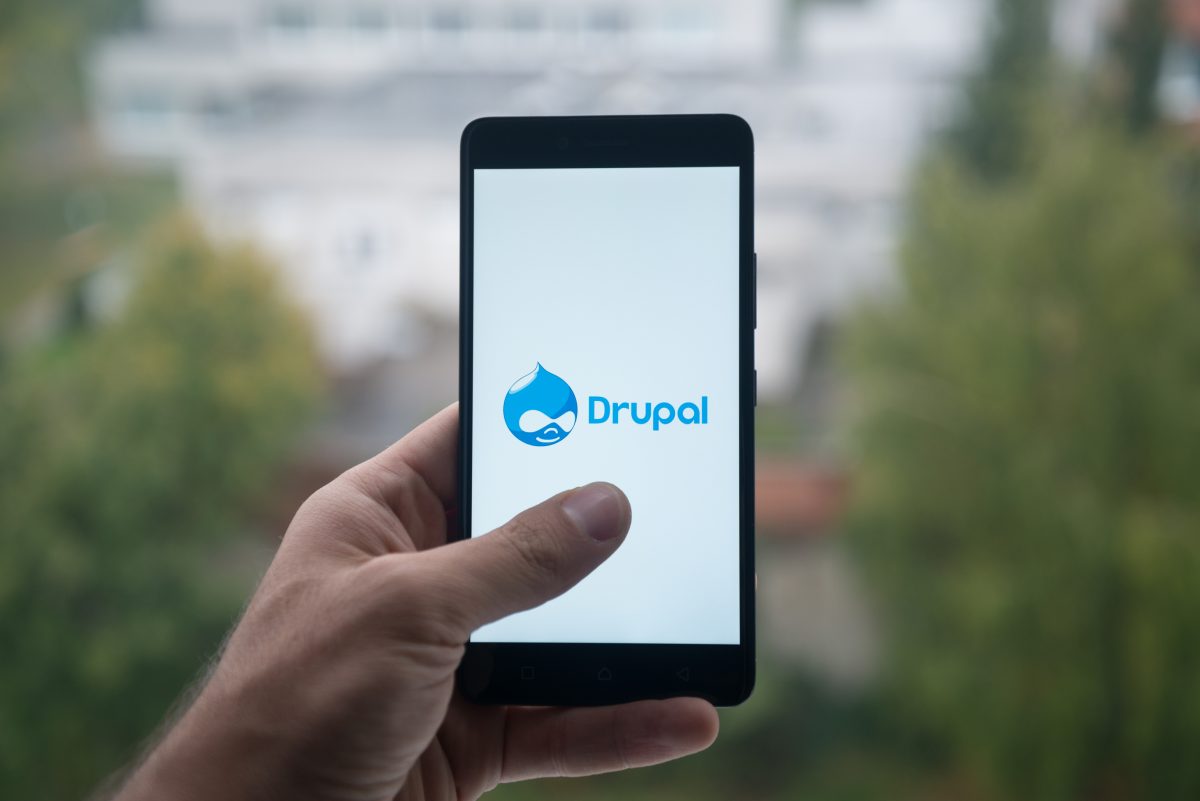 En hand håller en smartphone som visar Drupals logotyp mot en suddig grön utomhusbakgrund