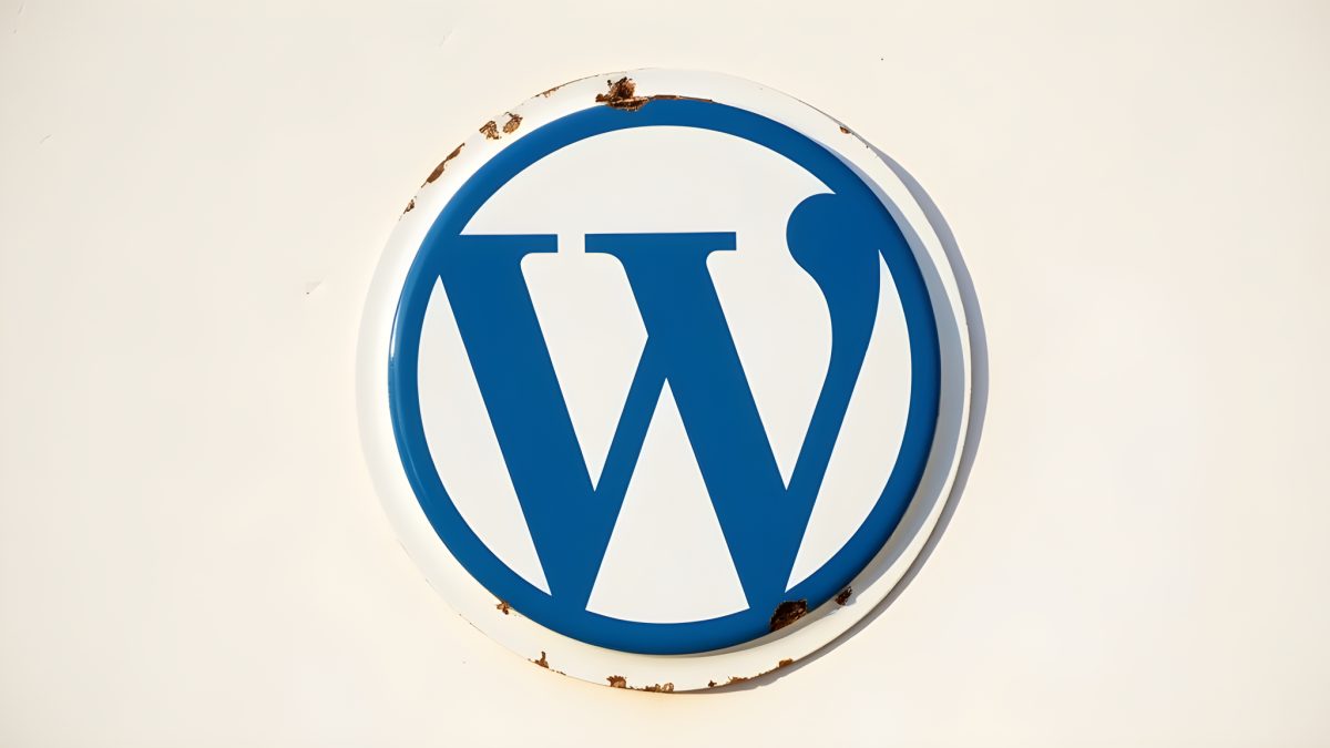 Rund emaljskylt med WordPress-logotypen i kobolt på vitt, med små rostflagor längs kanten, monterad på en solbelyst ljus vägg